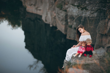 Young beautiful model-looking woman sitting on the rocks near the river with two dolls. A teenage girl imagines that she is a mother.