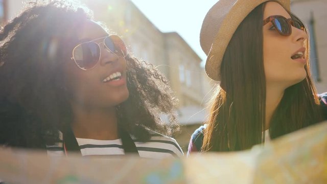 Two young pretty friends holding paper map in hands and standing in centre of old city. Multi-ethnic.
