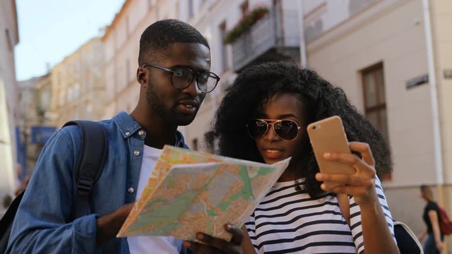 Tourists with city map and smartphone standing in centre of beautiful street. Young couple using internet and map for navigation.