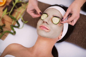 Spa. Beautiful young woman is getting facial clay mask at spa, lying with cucumbers on eyes
