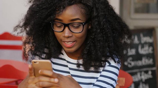 Young curly african american woman in glasses looking at mobile phone and typing message. Girl sitting at table in cafe. Close-up.