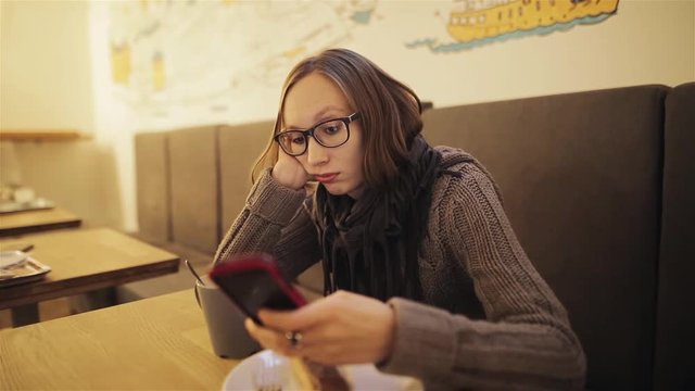A frustrated young impatient girl in glasses misses the cafe. The tired woman is waiting for the boyfriend and is bored with the phone in her hands. Love and relationships.