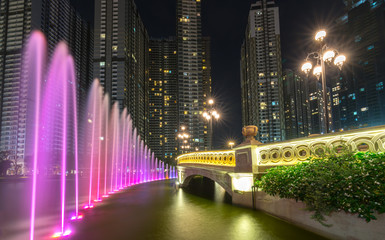 Fototapeta na wymiar Ho Chi Minh City, Vietnam - November 30th, 2017: A state of the art fountain at night with colorful lights shimmering, behind the skyscrapers in the urban park development in Ho Chi Minh City. Minh, V