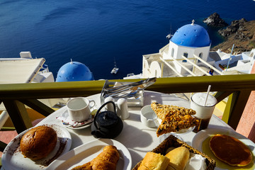 Breakfast with beautiful Aegean sea view, boat and Oia church including pancake, baguette, croissant, hot tea, chocolate brioche bun and fruity cereal bar on white marble table