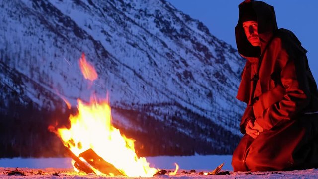 Alone monk in brown cloaks of the order of St. Francis is warming hands by the fire and prays