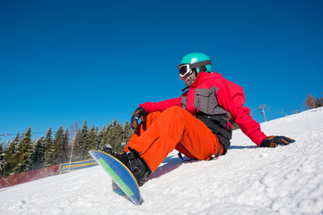 Male snowboarder sitting, relaxing on the slope of the hill at winter ski resort. Blue sky, forest...