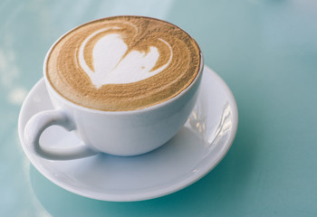 Cappuccino in white cup on green glass table, milk foam created as latte art in heart shape.