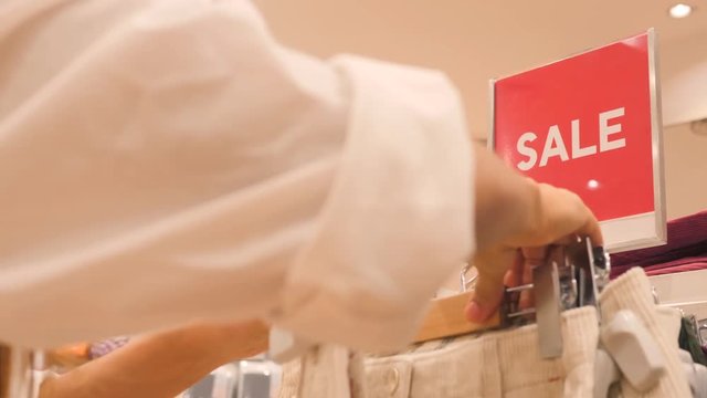 Young Woman Choosing Pants from Rack in Clothing Store. Big Red Sign in Shopping Mall. 4K.