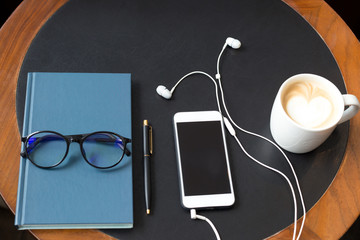 Cell phone with headphones and glasses on notebook and pen lying with coffee