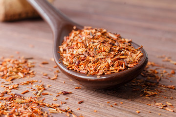 Composition of Rooibos tea is located on a ceramic spoon. Macro photo of a tea close-up. The petals of tea are located in a spoon on a wooden board. Natural ingredients of Rooibos tea.