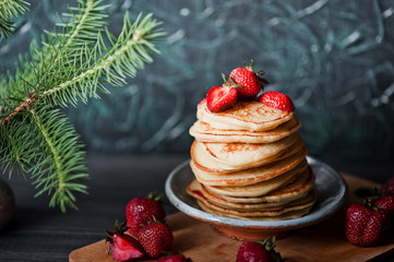 Stack of fritters with strawberries. Pancakes with red jam. Finished fritters on the background. Sprig of Christmas tree on the table.