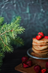 Stack of fritters with strawberries. Pancakes with red jam. Finished fritters on the background. Sprig of Christmas tree on the table.