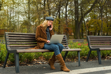 Beautiful young woman is sitting on the bench in the park on an autumn day and she is working on her laptop