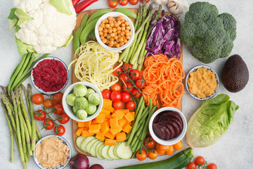 Above view of the ingredients for the salad, colourful vegetables, carrot, courgette, cabbage, chickpeas, cucumber and tomatoes, on the wooden board on white table, top view, selective focus