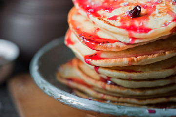 Stack of fritters with strawberries. Pancakes with red jam and coconut shavings. Finished fritters on the background. The man's hand pours pancakes with red jam. Sprig of Christmas tree on the table.