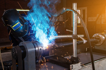 Close-up male auto mechanic in blue uniform in welding mask cooking metal on automatic welding...