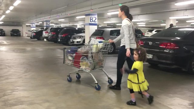 Mother and little daughter with a shopping cart in a parking lot after shopping activity in the supermarket. Consumption concept. Real people. Copy space