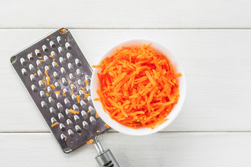 Grated fresh carrot in a white ceramic bowl and metal grater on a white wooden table top view