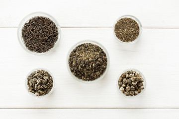 Different dry tea in a small glass dishes on a white wooden table, top view