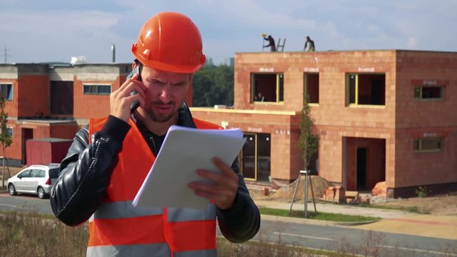 A young construction worker talks on a smartphone and looks at papers - a house under construction in the background