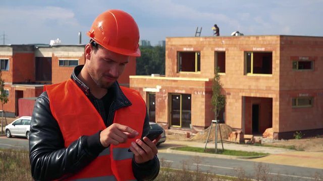A young construction worker works on a smartphone - a house under construction in the background
