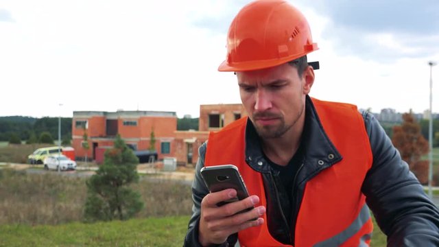 A young construction worker sits at a site and works on a smartphone - closeup - a house under construction in the background