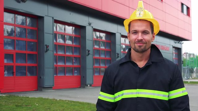 A young firefighter puts on a helmet and smiles at the camera with hands folded across his chest - a fire station in the background