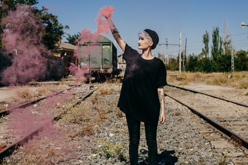 .Young modern tattoed woman dressed in black at an abandoned train station, playing with colorful smoke between train tracks. Lifestyle portrait