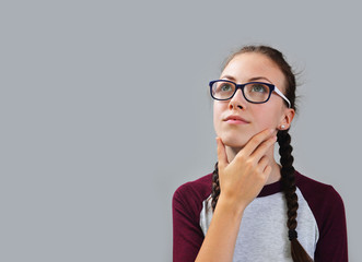 Young thoughtful girl with glasses dreaming on gray background.