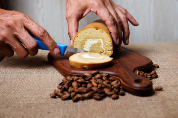 Man's hand is cutting  roll coffee cake with knife and coffee bean on wooden cutting board