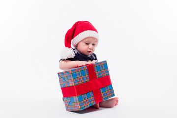 cute toddler is sitting in a New Year's red cap on white background unpacks a gift with a smile on his face