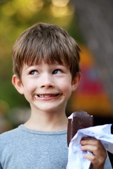 Happy boy in grey t shirt making faces while eating ice cream outdoor in the park. Close up funny facial expression, blurred background