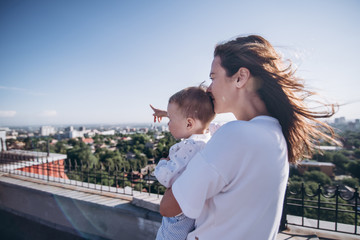 Beautiful brunette mother and little blonde son enjoy amazing roof top view of the city. Sunny morning, blu sky, old buildings, green trees and river on the horizont.