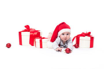 Cute baby in a red New Year's cap with a smile on his face lying on his stomach on a white background surrounded by boxes with gifts, and blue and red Christmas balls