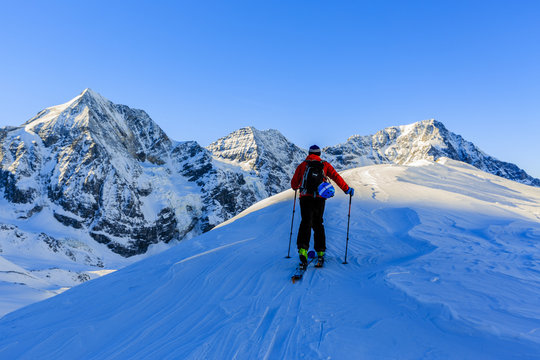 Mountaineer backcountry ski walking up along a snowy ridge with skis in the backpack. In background blue sky and shiny sun and Zebru, Ortler in South Tirol, Italy.  Adventure winter extreme sport.