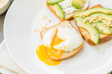 Breakfast. Eggs on toast with avocado, soft white cheese, spices and nuts. White plate, gray concrete background.