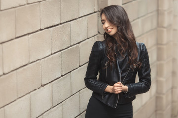 Portrait of beautiful smiling brunette, fashion and stylish standing near the wall. A curly hairstyle woman in total black look with leather jacket and dress.