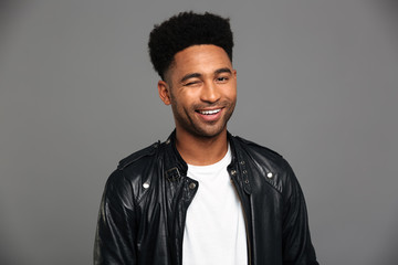 Fototapeta premium Young smiling african man in black leather jacket winks one eye, looking at camera