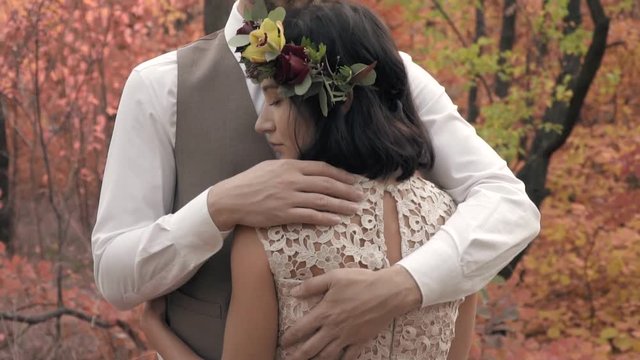 A slender woman in a wreath of leaves on her head hugs a man in a suit standing in the middle of the park in the autumn day