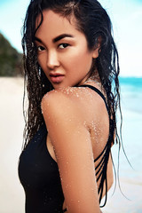 Fototapeta premium portrait of beautiful caucasian sunbathed woman model with dark long hair in dark swimsuit posing on summer beach with white sand on blue sky and ocean background