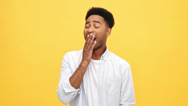 Tired african man in shirt yawns and covering his mouth over yellow background