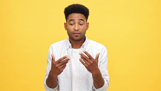 Confused african man in shirt shrugs his shoulders and looking at the camera over yellow background