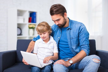 computer and internet concept - young father and son sitting on sofa with laptop