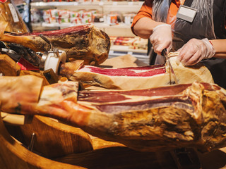 Hands of a woman in gloves in the market or store cut a slice of traditionally spanish jamon de bellota. Meat close up.