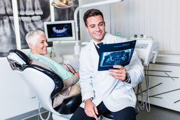 Handsome and attractive male dentist looking at dental x-ray together with his beautiful senior woman patient.