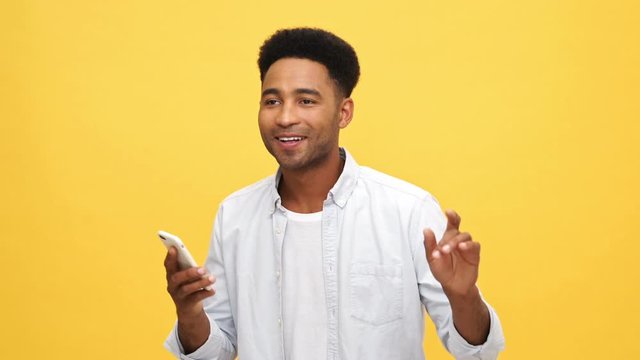 Smiling Young african man in shirt using his smartphone and listening music over yellow background
