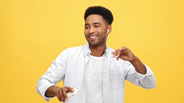 Happy Young african man in shirt listening music by earphones and dancing over yellow background