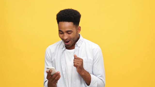 Happy surprised african man in shirt rejoice and looking at smartphone over yellow background