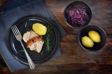 roast pork slices with rosemary on a plate, potatoes and red cabbage in bowls on a dark rustic wooden background, copy space, top view from above
