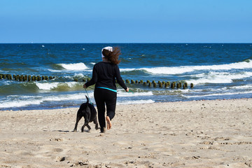 Kobieta spaceruje po plaży w wietrzny dzień z dużym czarnym psem. © Rajtar photography